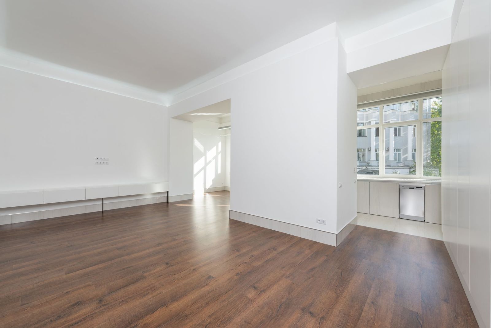 A bright empty apartment living space with wood floors and a kitchen beyond