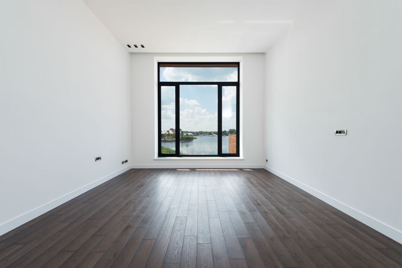 An empty room with dark wood floors and a tall window overlooking water