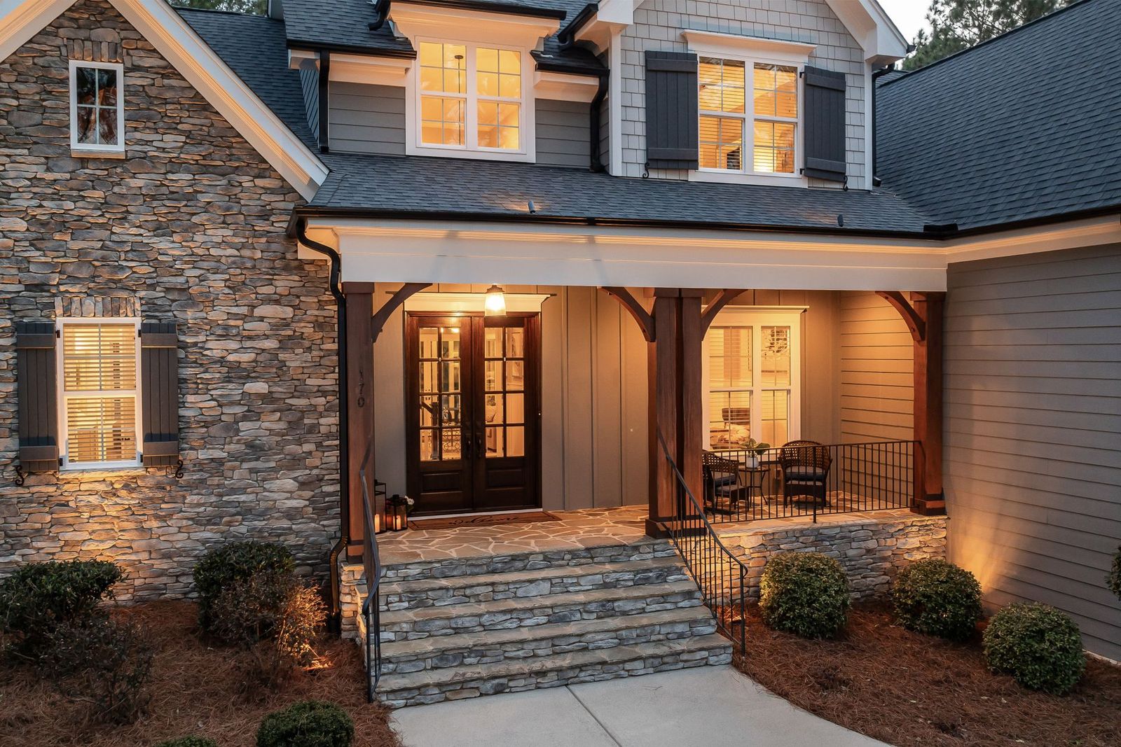 A stone-and-shingle house with its porch light and windows lit at dusk