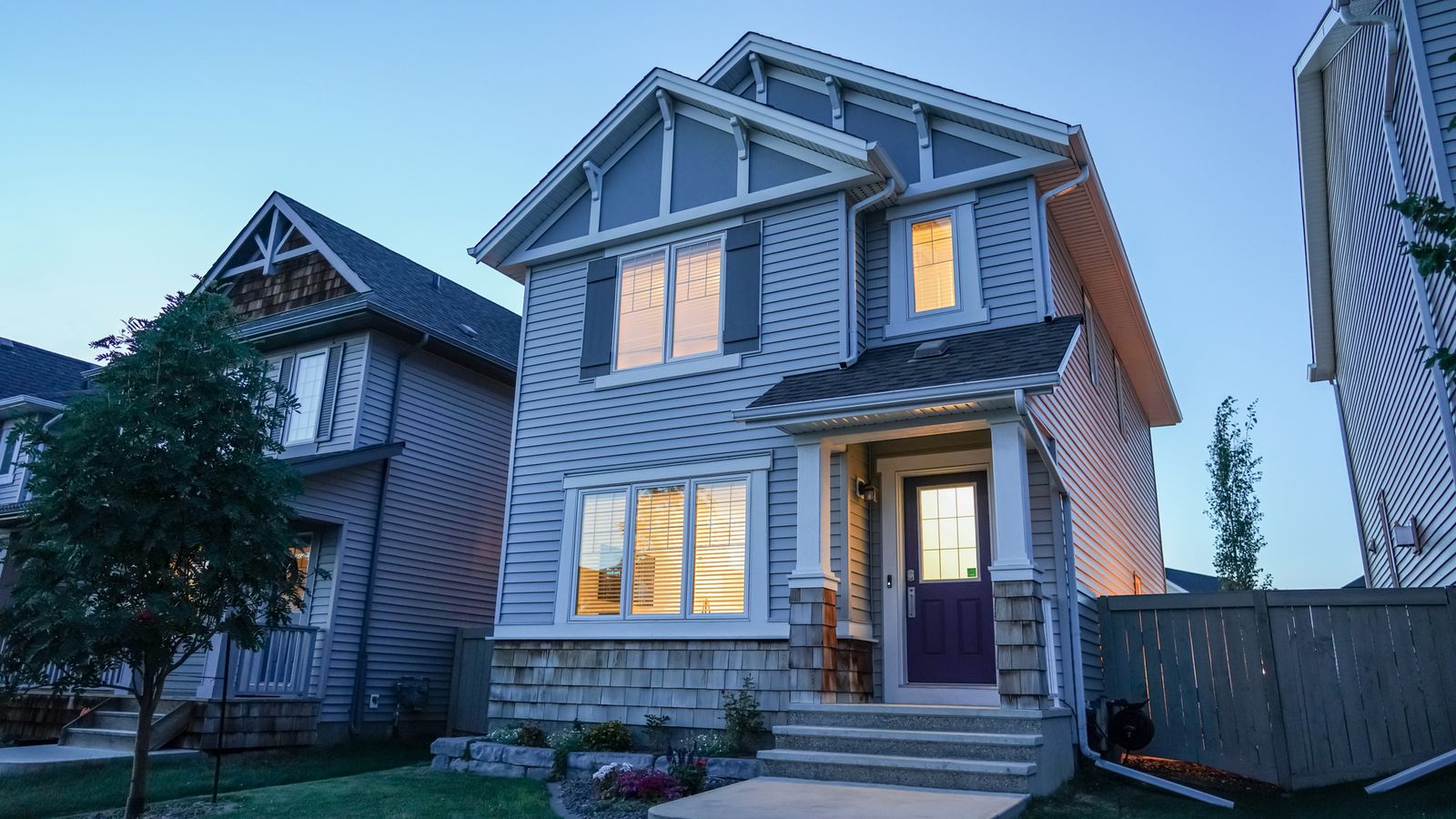 A two-story house with lit windows on a quiet suburban street at dusk